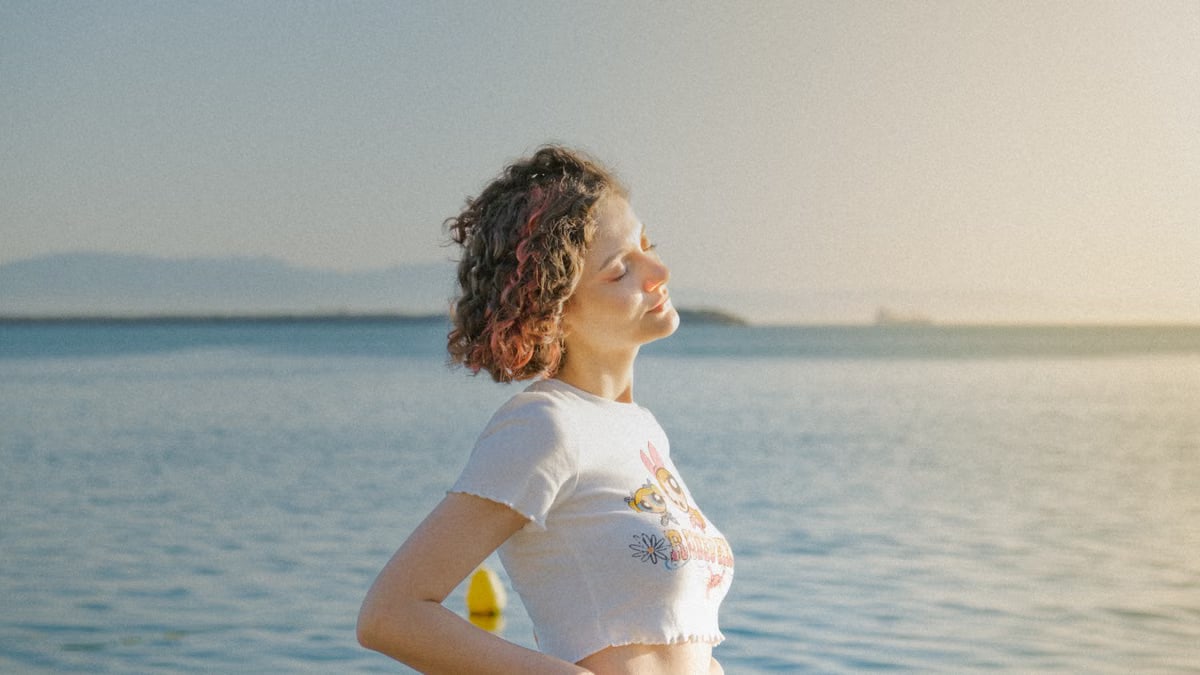 A woman with eyes closed stands by the ocean, embracing the sunlight and tranquility.